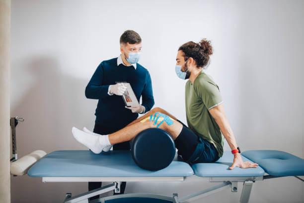 Athlete is sitting on the bed with pillow below his knees listening to the physical therapist explaining knee anatomy to him on the chart and what is a problem with his knee.