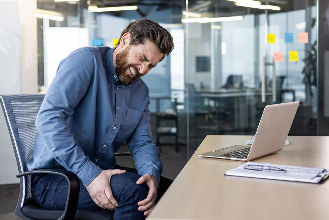 A young man sits at his desk in the office, grimacing in severe pain from a knee injury.